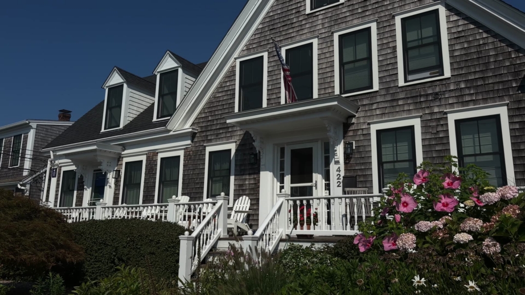 Front of a gray-shingled Cape Cod home with dormers, a white porch, and pink hibiscus