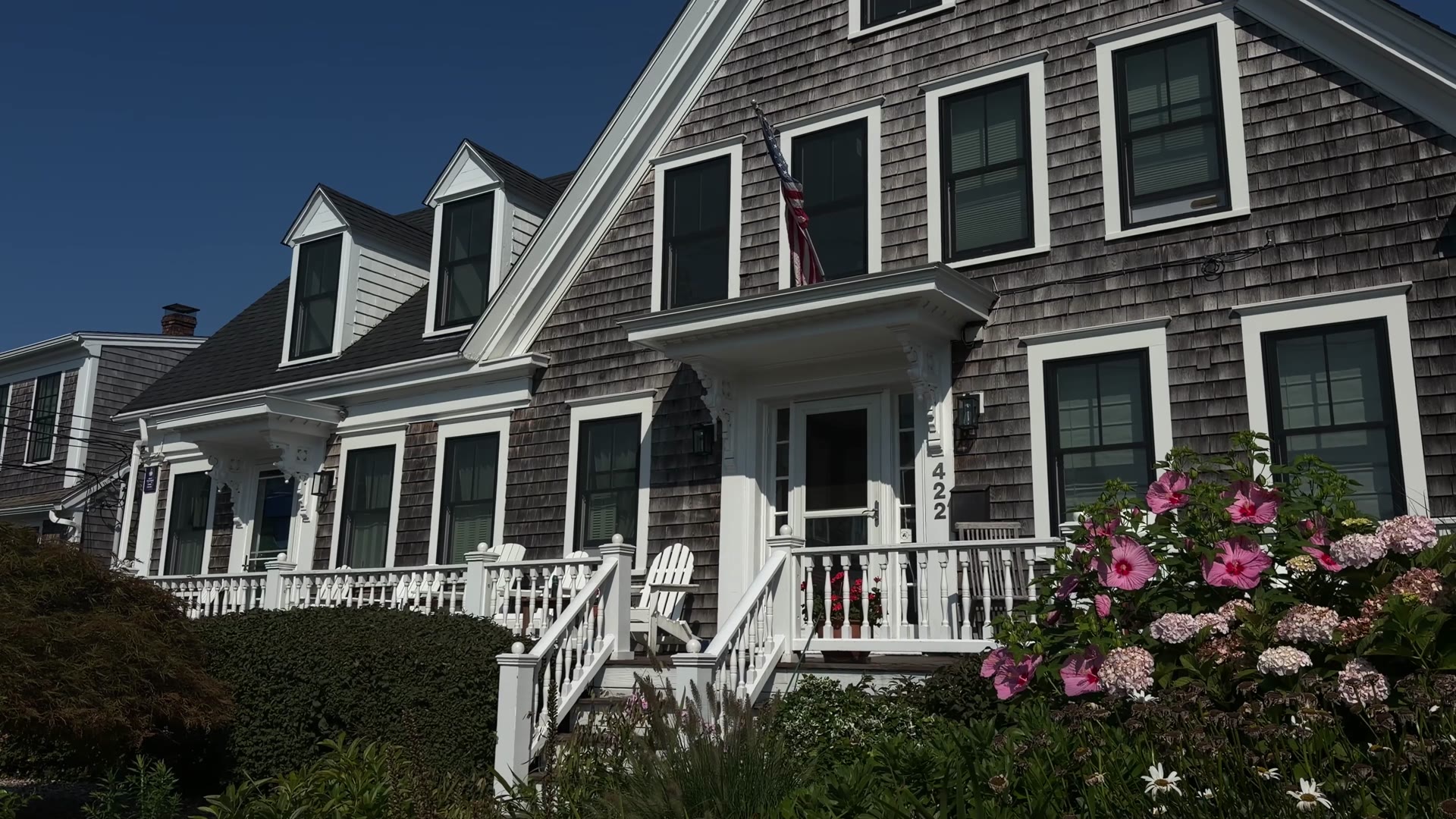 Front of a gray-shingled Cape Cod home with dormers, a white porch, and pink hibiscus