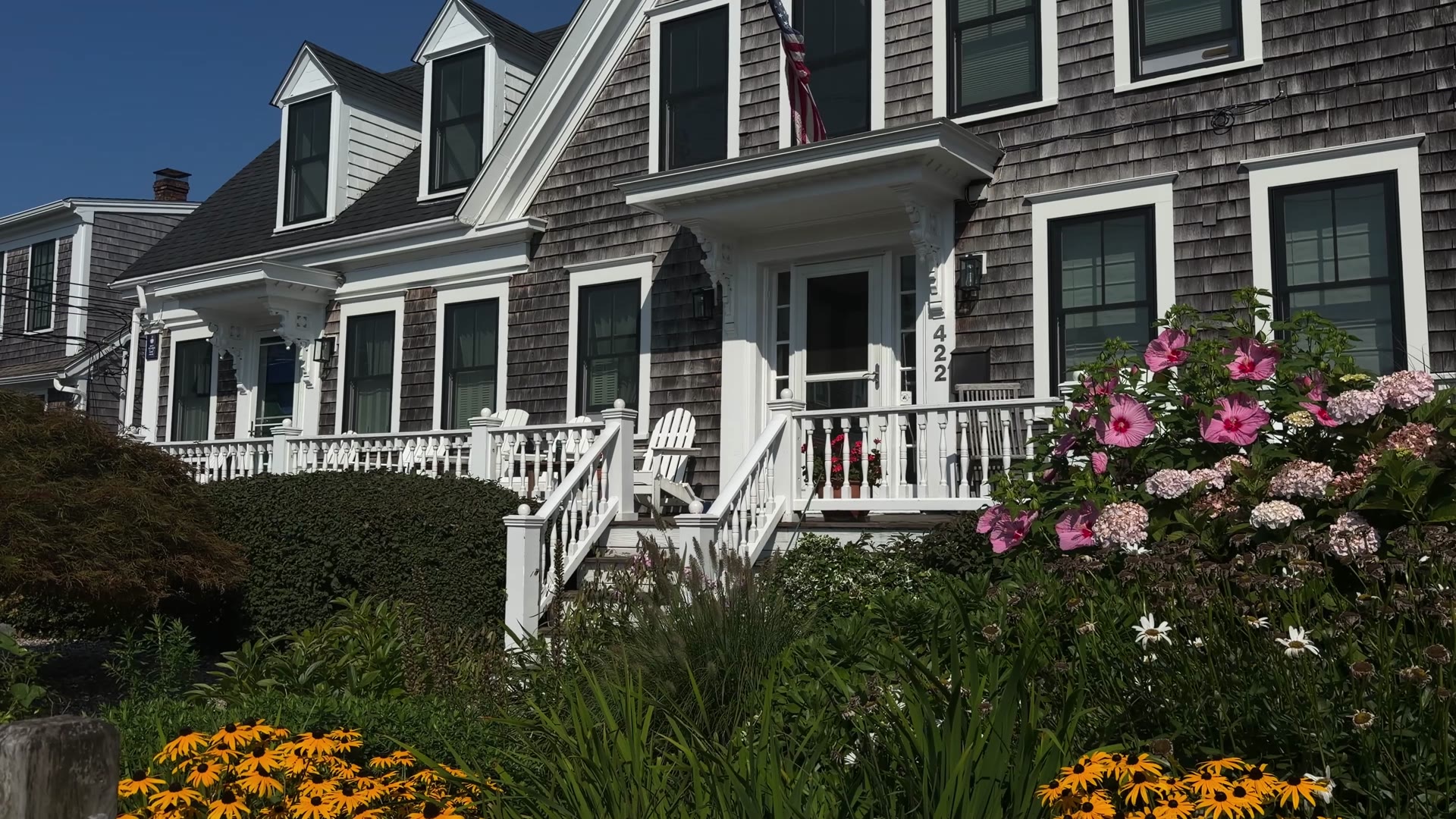 White porch, railings, and front steps of a gray-shingled Cape Cod home with flower gardens